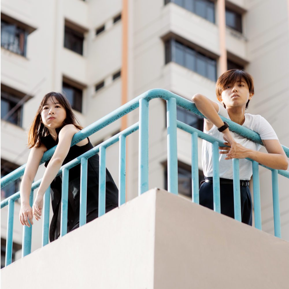 two young ladies standing on an outdoor terrace platform and resting on light blue metal railing in front of a large apartment complex